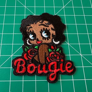 May include: An embroidered patch featuring a cartoon character with black hair, large blue eyes, and silver hoop earrings. The character is surrounded by red roses with green leaves. Below the character, the word Bougie is stitched in red thread.