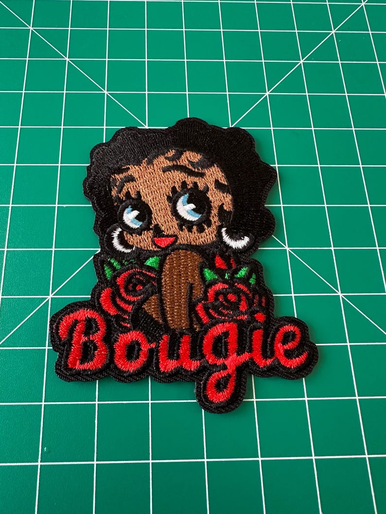 May include: An embroidered patch featuring a cartoon character with black hair, large blue eyes, and silver hoop earrings. The character is surrounded by red roses with green leaves. Below the character, the word Bougie is stitched in red thread.