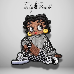 May include: An embroidered patch featuring a cartoon character with black hair, yellow hoop earrings, and red lips. The character wears a grey sweater and black and white checkered pants and sneakers. The text Truly Pressed is visible above.
