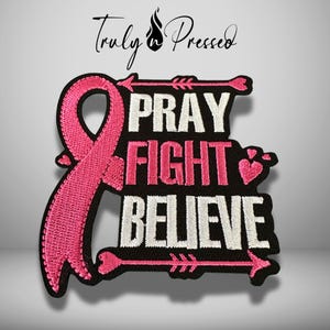 May include: A black embroidered patch featuring a pink awareness ribbon on the left side. The text PRAY FIGHT BELIEVE is stitched in white and pink. Two pink arrows point outward at the top and bottom, and three small pink hearts are included.