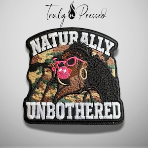 May include: An embroidered patch featuring a person with dark hair wearing pink sunglasses and blowing a pink bubble. The background is a camouflage pattern. The text reads NATURALLY UNBOTHERED in white block letters. The brand Truly n Pressed is above.