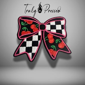 May include: An embroidered patch shaped like a bow with a black and white checkered pattern on two sections and a black background with red cherry illustrations on the other two sections, all outlined with a bright pink stitched border.