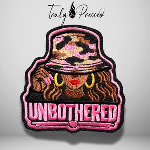 May include: An embroidered patch featuring a person wearing a pink, brown, and black camouflage bucket hat, gold hoop earrings, and red lipstick. The person has wavy brown hair. Below the figure, the word UNBOTHERED is stitched in pink.