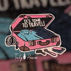 May include: Embroidered patch featuring a pink suitcase with the text "IT'S TIME TO TRAVEL!" The suitcase is open, revealing clothing inside. A small brush, spoon, and a stack of items are next to the suitcase. The patch has a white border.