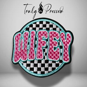 May include: Embroidered patch with the word "WIFEY" in pink, checkered letters, outlined in turquoise. The patch has a black border and a checkered background. The text "Truly & Pressed" is above the patch in black script.
