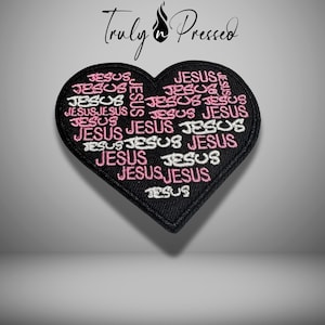May include: A black heart-shaped patch embroidered with the word "JESUS" in pink and white. The word is repeated multiple times, filling the heart shape. The patch has a black border and the text "Truly Pressed" is at the top.