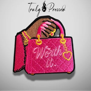 May include: An embroidered patch featuring a hand with dark skin tone holding a quilted hot pink handbag. The bag has gold hardware, a heart-shaped charm, and the text Worth it. embroidered in pink script on the front surface.