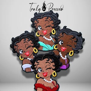 May include: An embroidered patch featuring four Betty Boop figures with black hair, gold hoop earrings, and white pearl necklaces. Each figure is winking with red lips. The figures wear dresses in teal, white, purple, and red. Text reads Truly Pressed.