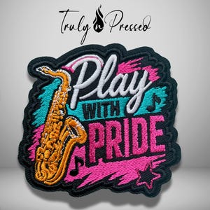 May include: An embroidered patch featuring a gold saxophone and the text "Play WITH PRIDE" in white and pink thread. The design includes teal and pink brushstroke accents, a black musical note, and a star on a black background with a black border.