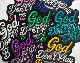 May include: A collection of embroidered fabric patches featuring the text God Don't Play Bout' Me in various thread colors including pink, blue, green, teal, purple, white, and yellow, all outlined with black stitching on a black background.