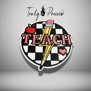 May include: A circular embroidered patch featuring a black and white checkered background. The word TEACH is stitched in pink letters across the center. A yellow pencil shaped like a lightning bolt, a red apple, and a small pink heart are included.