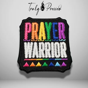 May include: A black embroidered patch with a scalloped edge featuring the text PRAYER in rainbow-colored thread and WARRIOR in white thread. Below the text is a horizontal rainbow line and a row of small, colorful embroidered triangles.