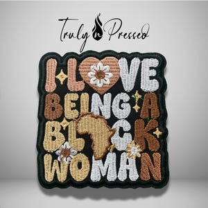 May include: A square embroidered patch with a black background featuring the text I LOVE BEING A BLACK WOMAN in various shades of brown, tan, and white. The letter O is replaced by a heart and a map of Africa, accented with small flower icons.