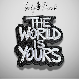 May include: A black embroidered patch featuring the white text THE WORLD IS YOURS in a bold, stylized font. The patch has a thick black border and a textured fabric base. Above the patch, the text Truly Pressed is printed in black.