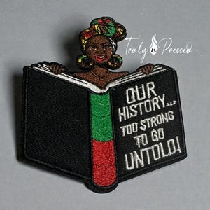 May include: Embroidered patch featuring a Black woman holding an open book. The book is black with a green and red spine and the text "OUR HISTORY... TOO STRONG TO GO UNTOLD!" The woman wears a colorful head wrap.