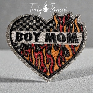 May include: Heart-shaped patch with a silver glitter border. The left side features a black and white checkered pattern, while the right side displays flames in red, orange, and yellow. The text "BOY MOM" is embroidered in silver.
