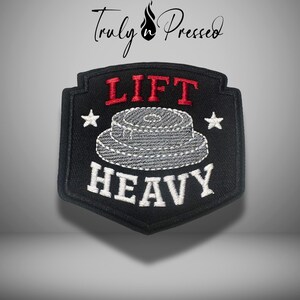 May include: Black embroidered patch with the words "LIFT HEAVY" in white and red. The patch features a weightlifting plate design in gray and two white stars. The text "Truly & Pressed" is at the top.