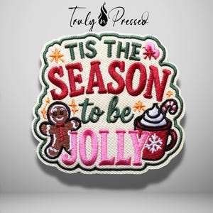 May include: An embroidered patch featuring the text TIS THE SEASON to be JOLLY in red, green, and pink thread. The design includes a brown gingerbread person, a red mug with white whipped cream and a candy cane, and small orange star accents.