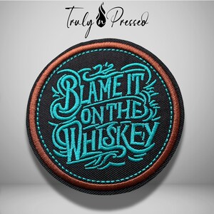 May include: A circular embroidered patch with a black background and a copper-colored border. The center features teal-colored decorative lettering that reads "BLAME IT ON THE WHISKEY" in a stylized script font with intricate swirling details.
