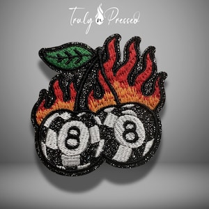 May include: An embroidered patch featuring two black eight balls with white number eight accents, surrounded by orange and red flames and topped with a single green leaf. The entire design is set against a black glittery background.