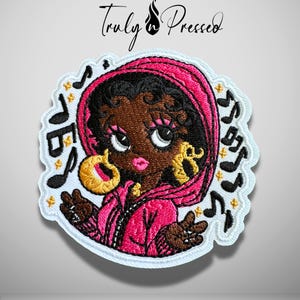 May include: An embroidered patch featuring a cartoon character with dark skin, black hair, and large eyes, wearing a bright pink hooded jacket and gold hoop earrings. The design includes black musical notes and small yellow stars on a white background, with the text Truly Pressed at the top.
