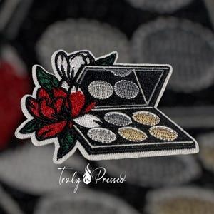 May include: Embroidered patch featuring a black eyeshadow palette with silver and gold shades, accented by a red rose and white flower. The patch has a white border and the text "Truly Pressed" in cursive.
