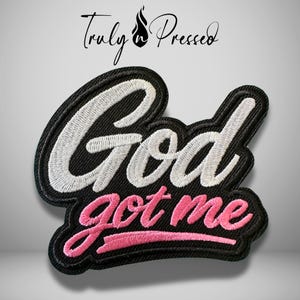 May include: Embroidered patch with the words "God got me" in white and pink thread on a black background. The patch has a black border and the text "Truly Pressed" in cursive.