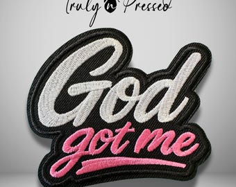 May include: An embroidered patch with a black background and a thick black border. The text God is stitched in white thread in a cursive script, while the text got me is stitched in pink thread below it, also in a cursive script.