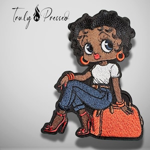 May include: An embroidered patch featuring a cartoon character with black hair, wearing a white t-shirt, blue jeans, and red high heels, sitting on an orange bag. The text Truly n Pressed is visible in the upper left corner of the image.