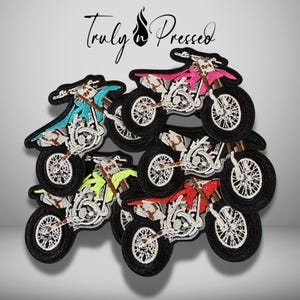 May include: A collection of embroidered motorcycle patches featuring the text Truly n Pressed at the top. The patches depict dirt bikes in various colors including pink, teal, neon yellow, and red, all with black tires and white engine details.