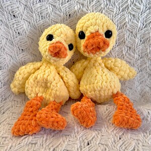May include: Two handmade yellow crochet duck toys with orange beaks and feet. The ducks have black eyes and are made with a soft, textured yarn. The ducks are sitting on a white, textured surface.