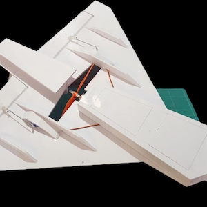 Peut inclure: Un mod&egrave;le r&eacute;duit d'avion blanc avec une aile delta. Le mod&egrave;le a un long corps rectangulaire et plusieurs petites ailes. L'h&eacute;lice est orange et noire. Le mod&egrave;le est sur un tapis de coupe vert.