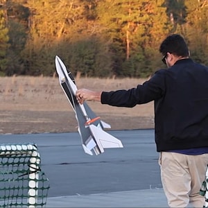 Peut inclure: Une personne tient un mod&egrave;le d'avion &agrave; l'ext&eacute;rieur. L'avion est blanc, noir et gris avec des accents orange. L'arri&egrave;re-plan comprend des arbres et une zone pav&eacute;e.