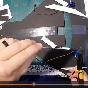 Peut inclure: Une personne travaille sur un mod&egrave;le d'avion. Le mod&egrave;le est fait de pi&egrave;ces de mousse noires et blanches. La personne utilise une pince et d'autres outils pour assembler l'avion. L'arri&egrave;re-plan est un tapis de coupe vert.