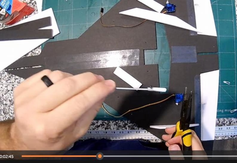 Peut inclure: Une personne travaille sur un mod&egrave;le d'avion. Le mod&egrave;le est fait de pi&egrave;ces de mousse noires et blanches. La personne utilise une pince et d'autres outils pour assembler l'avion. L'arri&egrave;re-plan est un tapis de coupe vert.