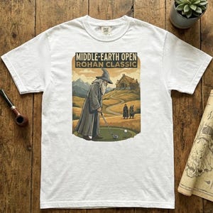 May include: White t-shirt featuring a graphic of a wizard playing golf with the text "MIDDLE-EARTH OPEN ROHAN CLASSIC". The design has a vintage poster style with a golf course and mountain backdrop.