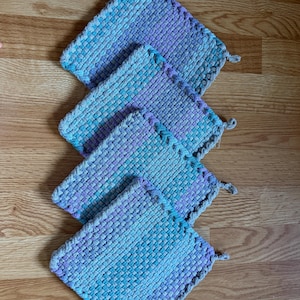 May include: Four square potholders arranged in a diagonal line. Each potholder features a woven texture with a gradient pattern transitioning from light blue to purple and grey. Each piece includes a small loop on one corner for hanging.