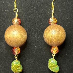 May include: A pair of dangle earrings featuring gold-tone metal hooks, a small amber-colored crackle glass bead, a large round wooden bead with a visible grain, another amber crackle glass bead, and a small green owl-shaped charm at the base.