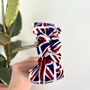 Puede incluir: Una diadema blanca con un patr&oacute;n de Union Jack rojo, blanco y azul. La diadema est&aacute; anudada en la parte superior.