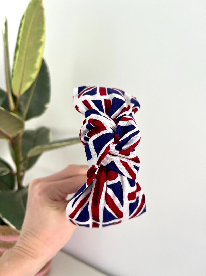 Puede incluir: Una diadema blanca con un patr&oacute;n de Union Jack rojo, blanco y azul. La diadema est&aacute; anudada en la parte superior.