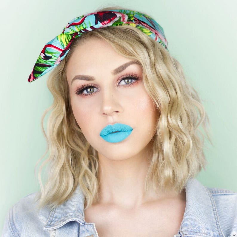 Puede incluir: Una mujer con el pelo rubio y ondulado lleva una chaqueta vaquera azul y una diadema con estampado tropical. Lleva un pintalabios azul brillante.