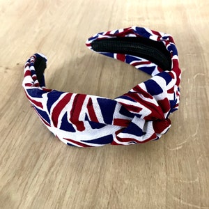 Puede incluir: Una diadema blanca, azul y roja con un dise&ntilde;o anudado. La diadema est&aacute; hecha de tela y tiene un patr&oacute;n de Union Jack.