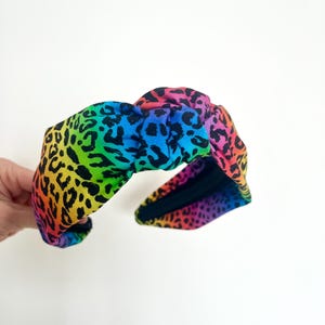Könnte beinhalten: Ein Stirnband mit Regenbogen-Leopardenmuster. Das Stirnband hat oben einen Knoten und eine schwarze Innenseite. Die leuchtenden Farben gehen von Gelb über Grün, Blau und Lila bis hin zu schwarzen Flecken.