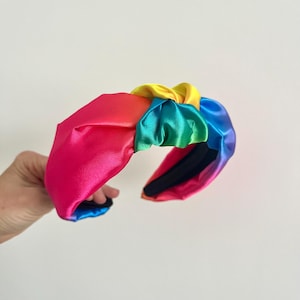 Könnte beinhalten: Ein farbenfrohes Satin-Haarband in Regenbogenfarben. Das Haarband hat ein geknotetes Design mit einem Farbverlauf aus Pink, Rot, Gelb, Grün und Blau. Das Haarband wird von einer Hand vor einem weißen Hintergrund gehalten.
