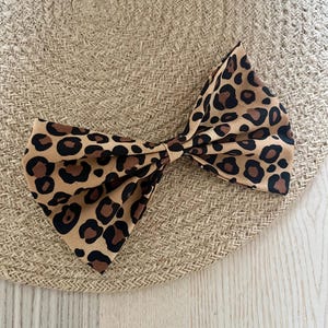 Peut inclure: Un chapeau de soleil beige avec un grand nœud. Le nœud présente un fond beige avec des taches de léopard marron et noir. Le chapeau a une texture tissée. Le chapeau et le nœud sont sur une surface en bois clair.