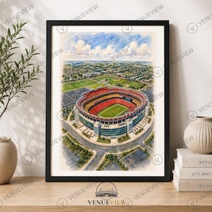 May include: A framed watercolour-style art print depicting an aerial view of a large circular sports stadium with red seating, a green field, and surrounding parking lots under a blue sky with white clouds, presented in a black rectangular frame.
