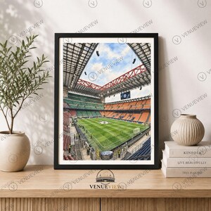 May include: A framed wall art print depicting a high-angle view of a large soccer stadium with a green grass pitch, orange and green seating sections, a metal roof structure, and a blue sky visible through the open center of the stadium roof.