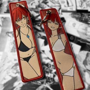 May include: Two rectangular keychains with embroidered anime-style illustrations of a woman in a bikini. The keychains have a red border and a silver key ring. One bikini is black, the other is white.