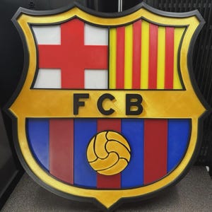 May include: A wall-mounted FC Barcelona crest emblem featuring a black border, a white section with a red cross, a yellow section with red vertical stripes, and a lower section with red and blue stripes containing a central yellow football.