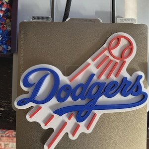 Può includere: Un logo dei Dodgers stampato in 3D in blu, bianco e rosso. Il logo presenta la scritta "Dodgers" in blu, uno sfondo bianco e una palla da baseball con linee rosse. Il logo è posizionato su una superficie metallica strutturata.